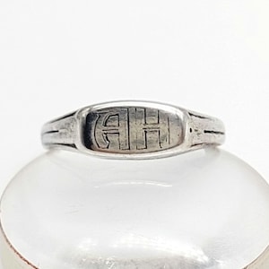 Può includere: Un anello con sigillo d'argento con le iniziali incise "AH" in un'impostazione rettangolare. L'anello ha una fascia arrotondata e una faccia leggermente rialzata. L'anello è esposto su una superficie trasparente e arrotondata.