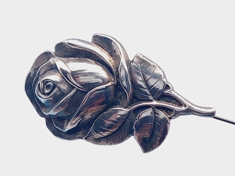 May include: A silver rose brooch with a detailed design of petals and leaves. The brooch has a pin on the back for attaching to clothing.