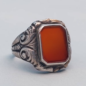 Könnte beinhalten: Ein silberner Ring mit einem verzierten Design und einem großen, rechteckigen, orangefarbenen Karneol-Stein.
