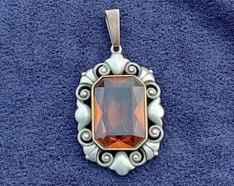 May include: A silver pendant with an ornate design featuring a large, rectangular, orange-brown gemstone.