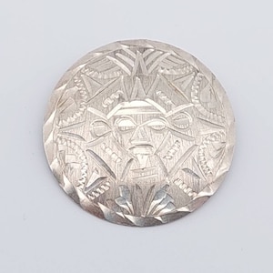 Puede incluir: Un broche de plata con un diseño detallado de una cara rodeada de patrones geométricos.