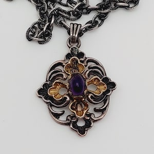 Puede incluir: Un collar de plata y oro con un colgante que tiene una piedra preciosa p&uacute;rpura en el centro. El colgante tiene forma de cruz con intrincados dise&ntilde;os florales.