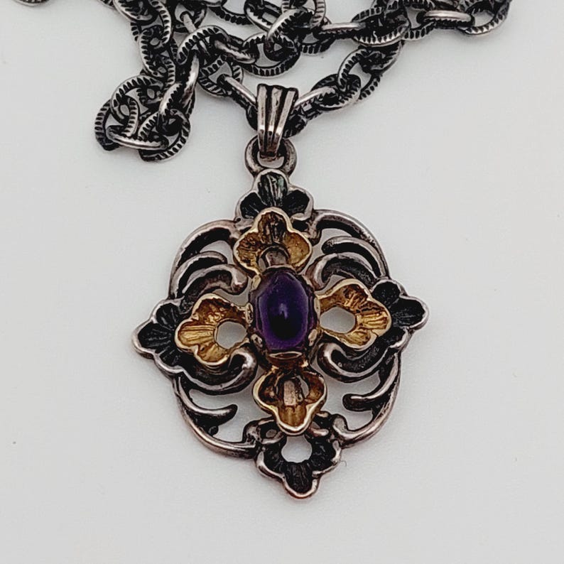 Puede incluir: Un collar de plata y oro con un colgante que tiene una piedra preciosa p&uacute;rpura en el centro. El colgante tiene forma de cruz con intrincados dise&ntilde;os florales.