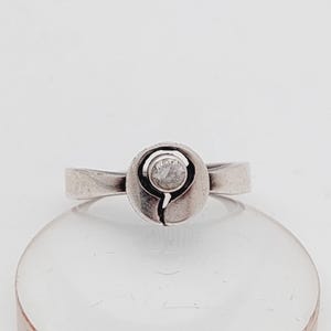 Puede incluir: Un anillo de plata con un diseño redondo calado que presenta una sola piedra preciosa transparente.