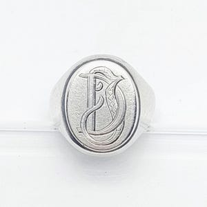 May include: A silver-toned signet ring featuring an oval face with a polished finish. The center of the ring is engraved with a large, ornate, stylized letter D in a decorative script font, showcasing intricate line work and detailing.