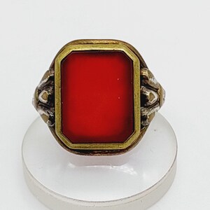 May include: Vintage ring with a rectangular, bright red stone set in a gold-toned frame. The ring has ornate, silver-toned detailing on the sides. The ring is displayed on a white, circular stand.