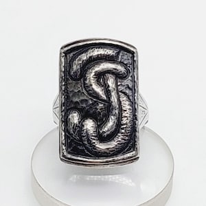 May include: Silver ring with a rectangular face. The face has a raised, textured design of a stylized snake. The ring has a hammered metal appearance. It is displayed on a clear stand.