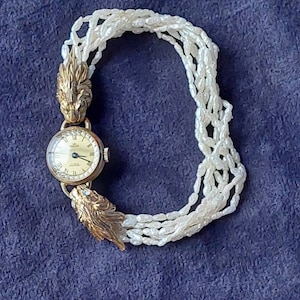 May include: A gold wristwatch with a white face and black hands. The watch is attached to a white pearl bracelet with gold lion head clasps.