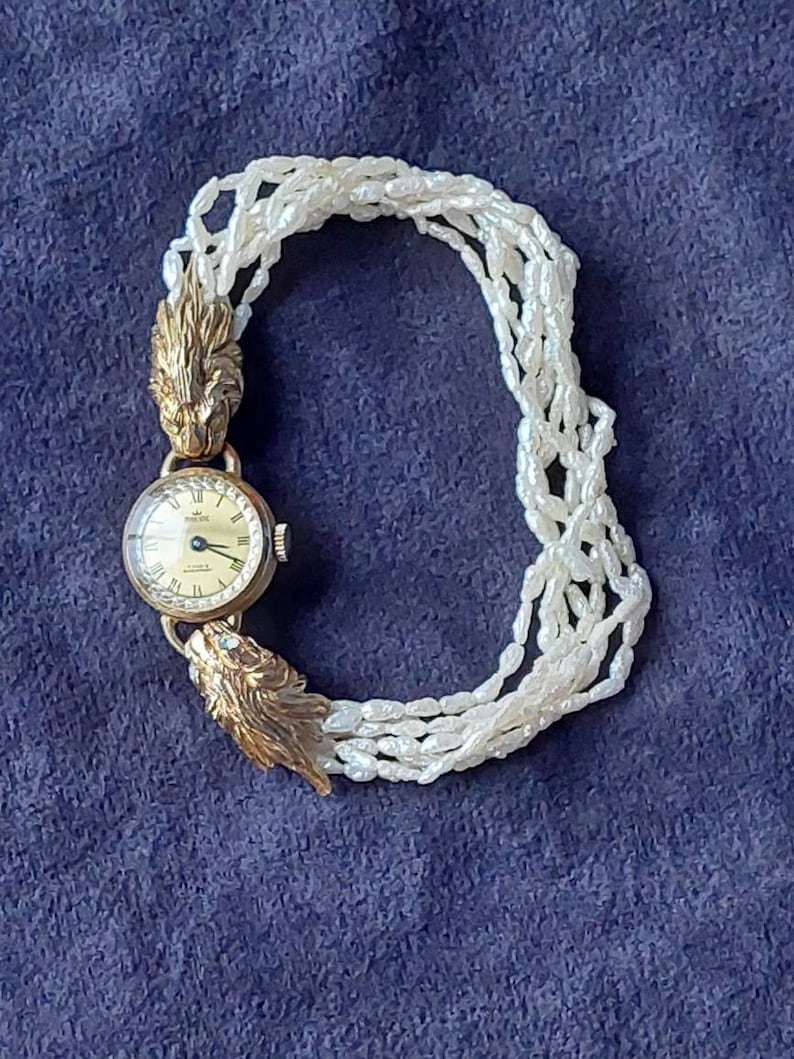 May include: A gold wristwatch with a white face and black hands. The watch is attached to a white pearl bracelet with gold lion head clasps.