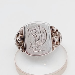 Op de afbeelding: Een zilveren zegelring met een gegraveerd rechthoekig vlak. Het vlak heeft een sierlijke, gestileerde letter "D". De ring heeft decoratief krulwerk op de schouders. De ring wordt getoond op een heldere, ronde standaard.