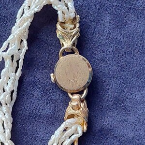May include: A gold-tone wristwatch with a pearl bracelet. The watch face is covered with a glass dome and the back of the watch has the number "0333" engraved on it.
