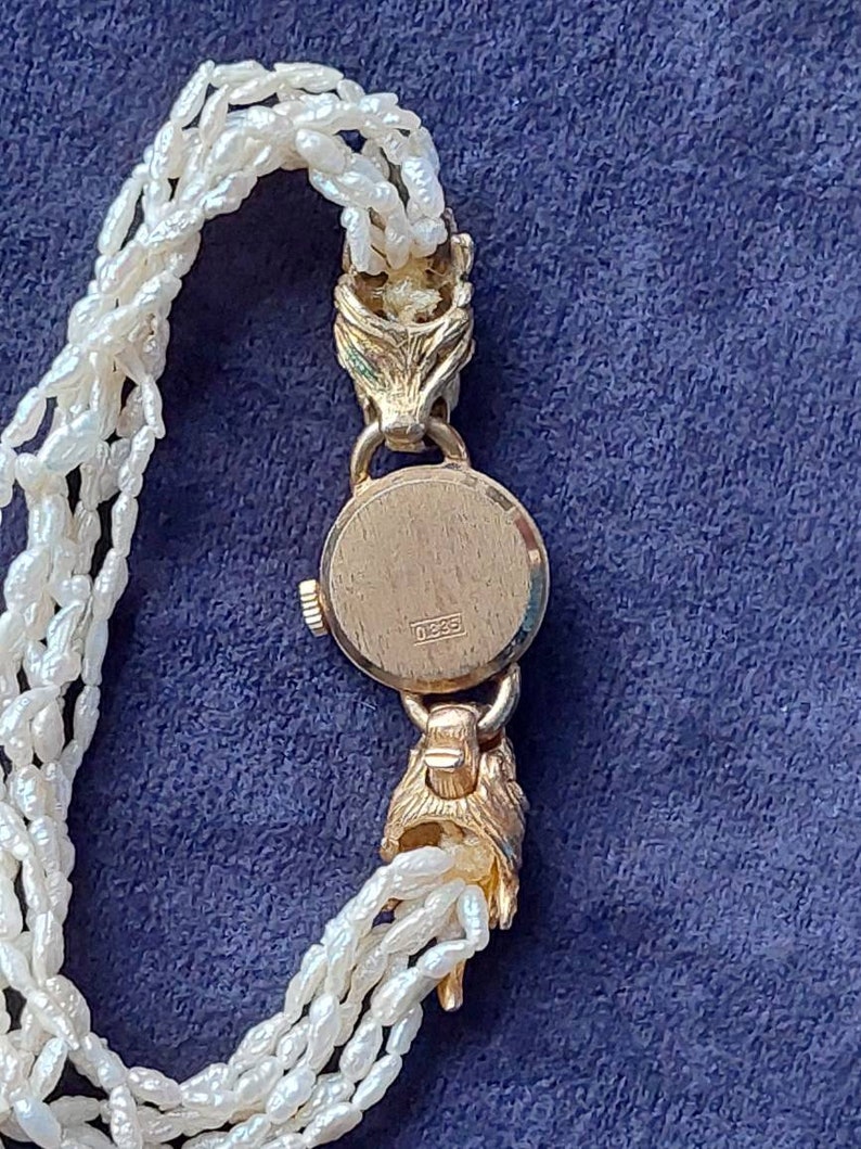 May include: A gold-tone wristwatch with a pearl bracelet. The watch face is covered with a glass dome and the back of the watch has the number "0333" engraved on it.