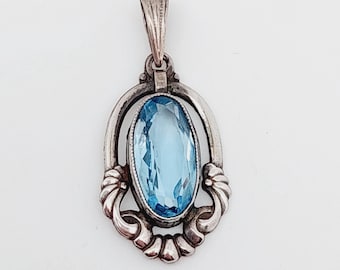 May include: A silver pendant with an oval, light blue gemstone. The pendant has an ornate, vintage design with decorative flourishes around the gemstone. The bail is a simple, textured loop.