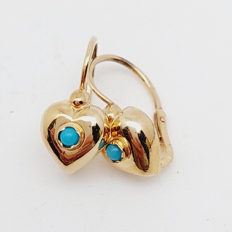 May include: A pair of gold-colored heart-shaped earrings with a small turquoise stone in the center of each heart. The earrings have a lever back closure and are displayed against a white background.