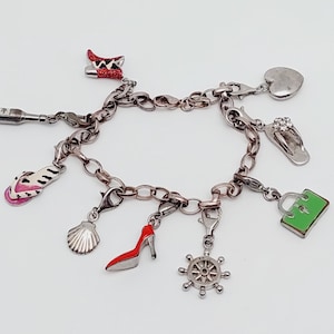 Puede incluir: Pulsera de plata con varios dijes, incluyendo un zapato de tacón, una chancla, una concha, un bolso, un corazón, un timón y una botella. Los dijes son plateados, rojos, rosas y verdes.