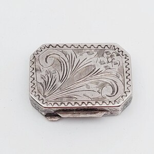 May include: A rectangular silver box with intricate floral and leaf engravings. The box has a zig-zag border and a hinged lid. The detailed design covers the top surface, showcasing a vintage aesthetic.