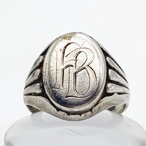 May include: A silver signet ring with an oval face. The face displays the intertwined letters "HB" in a decorative font. The ring has a ribbed design on the sides and a slightly worn look, suggesting it is vintage.