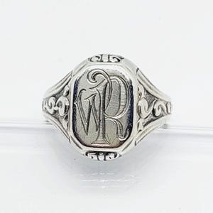 Può includere: Un anello sigillo in argento con una faccia ovale caratterizzata da un monogramma inciso delle lettere W e P in corsivo. La fascia e i lati dell'anello sono decorati con intricati motivi a volute e disegni in rilievo con finitura metallica.