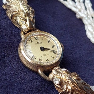May include: A gold-tone wristwatch with a white face and black hands. The watch has a lion head design on each side of the face. The watch is on a white pearl necklace.