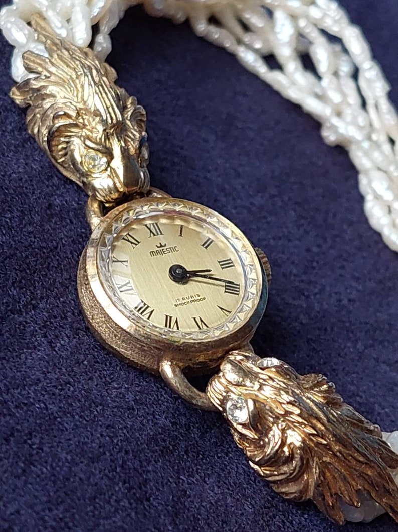 May include: A gold-tone wristwatch with a white face and black hands. The watch has a lion head design on each side of the face. The watch is on a white pearl necklace.