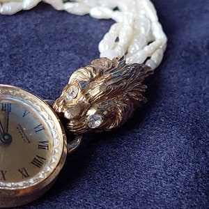 May include: A gold-tone wristwatch with a white face and black hands. The watch has Roman numeral hour markers and a lion head design on the band. The band is made of white pearls.