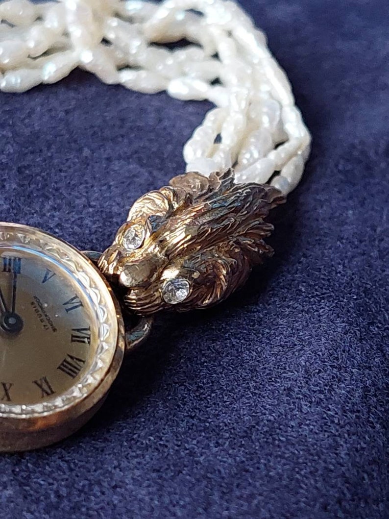 May include: A gold-tone wristwatch with a white face and black hands. The watch has Roman numeral hour markers and a lion head design on the band. The band is made of white pearls.