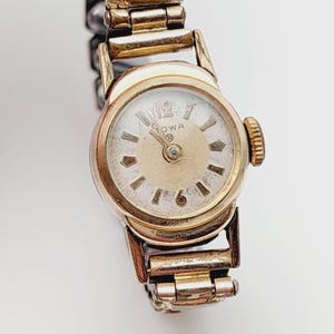 May include: A gold-toned wristwatch with a white face and black Roman numeral hour markers. The watch has a gold-toned metal band and says "ROWA" on the face.