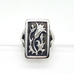 May include: A rectangular silver ring featuring a raised floral motif set against a dark, oxidised background. The central design depicts a single flower with detailed petals and leaves. The band of the ring has decorative side accents.