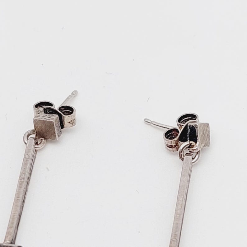 Puede incluir: Un par de pendientes de plata con un colgante cuadrado en la parte inferior. Los pendientes tienen un dise&ntilde;o simple y minimalista.