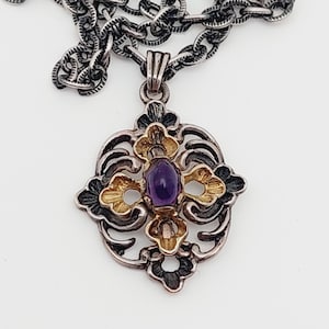 Puede incluir: Un collar de plata y oro con un colgante que tiene una piedra preciosa p&uacute;rpura en el centro. El colgante tiene forma de cruz con intrincados dise&ntilde;os florales.