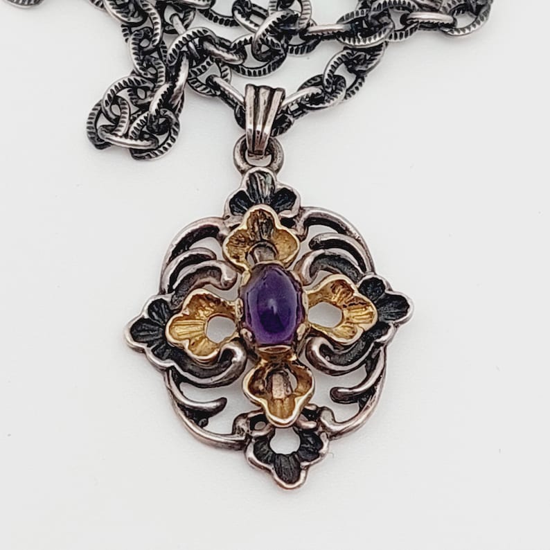 Puede incluir: Un collar de plata y oro con un colgante que tiene una piedra preciosa p&uacute;rpura en el centro. El colgante tiene forma de cruz con intrincados dise&ntilde;os florales.