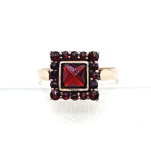Könnte beinhalten: Ein goldfarbener Ring mit einem quadratischen, tiefroten Edelstein in der Mitte, umgeben von kleineren, passenden roten Steinen. Das Design des Rings ist symmetrisch, mit einer klassischen, Vintage-Ästhetik. Das Band ist schlichtes, poliertes Gold.