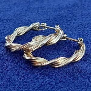 Puede incluir: Un par de pendientes de aro retorcidos en oro y plata. Los pendientes son de metal y tienen un acabado pulido.