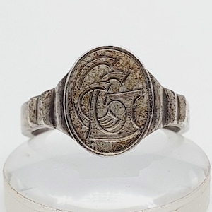 Puede incluir: Un anillo de sello plateado con una cara ovalada. La cara presenta un diseño grabado de letras entrelazadas. La banda tiene un diseño estriado en los lados. El anillo se muestra sobre un soporte transparente sobre un fondo blanco.