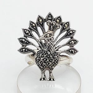 May include: A silver ring with a peacock design. The peacock's body and tail feathers are embellished with small, dark stones, giving a detailed and textured look. The ring is set against a white backdrop.