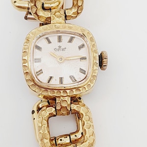 May include: A gold-toned wristwatch with a square face and a textured gold band. The watch face has black Roman numeral hour markers and a white background with the word "Tissot" printed in black.