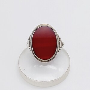 May include: A silver ring featuring a large, oval, deep red stone. The stone is set in a silver band with decorative detailing on the sides. The ring is displayed on a clear, round stand against a white background.
