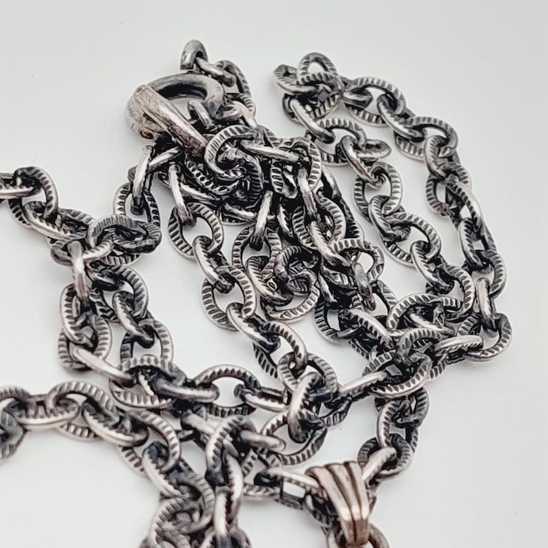 Puede incluir: Un collar de cadena de plata con un dise&ntilde;o de eslab&oacute;n texturizado de forma ovalada. La cadena tiene un cierre de mosquet&oacute;n.