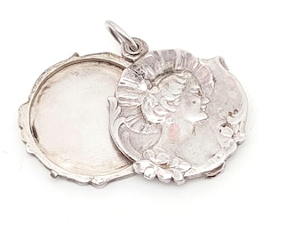 May include: A silver-colored locket with an oval shape, open to reveal a blank interior. The front features a detailed relief of a woman's profile, adorned with flowers and a decorative border. The locket has a small loop for a chain.
