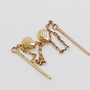 Puede incluir: Un par de pendientes de oro con una cadena delicada y un diseño de concha. Los pendientes están hechos de oro y tienen un diseño simple y elegante.