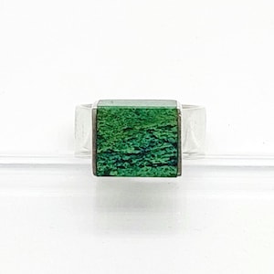 Peut inclure: Une bague en argent dotée d'une incrustation carrée à la surface texturée d'un vert éclatant. L'anneau en métal est lisse et poli, encadrant le carré central vert qui présente une finition brute et naturelle aux tons émeraude profonds.
