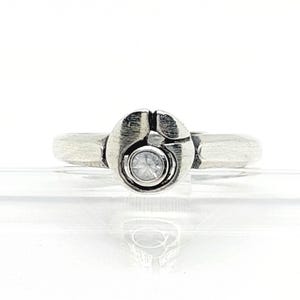 Puede incluir: Un anillo de tono plateado con acabado de metal cepillado y una pieza central circular. El centro del anillo contiene una pequeña piedra preciosa transparente de corte redondo, engastada en un marco circular hundido con una hendidura vertical.