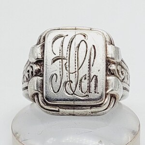 Könnte beinhalten: Ein silberner Ring mit rechteckiger Oberfläche, auf der die Initialen "Jch" in einer eleganten Schrift eingraviert sind. Der Ring hat ein detailliertes, verziertes Design an den Seiten und ein leicht gealtertes Aussehen, was auf Vintage oder Antike hindeutet.