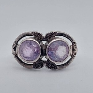 May include: A silver ring with two round, light purple gemstones set in a delicate, floral design.