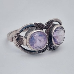 May include: A silver ring with two light purple gemstones set in a delicate, ornate design.