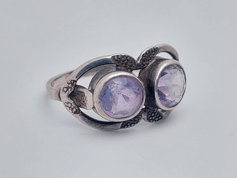 May include: A silver ring with two light purple gemstones set in a delicate, ornate design.