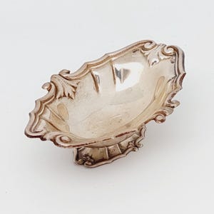 May include: A small, silver-coloured dish with an ornate, scalloped edge. The dish has a shallow, oval bowl and a decorative base. The design includes floral and scrollwork details, giving it an antique or vintage appearance. The dish is likely made of metal.