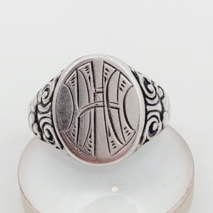 Può includere: Anello con sigillo argentato con una faccia ovale con una lettera "H" stilizzata e ornata. L'anello presenta un motivo a spirale dettagliato attorno alla fascia. L'anello è presentato su una superficie bianca.
