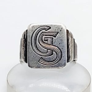 May include: A silver-toned signet ring with a rectangular face. The face features the intertwined letters "C", "G", and "S" in a bold, stylized font. The ring has a thick band and a slightly aged appearance.