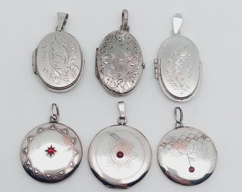 May include: Six silver lockets with different designs. Three are oval, with floral and geometric patterns. The other three are round, with starburst, floral, and geometric patterns. Each locket has a loop for a chain and some have a red gem.